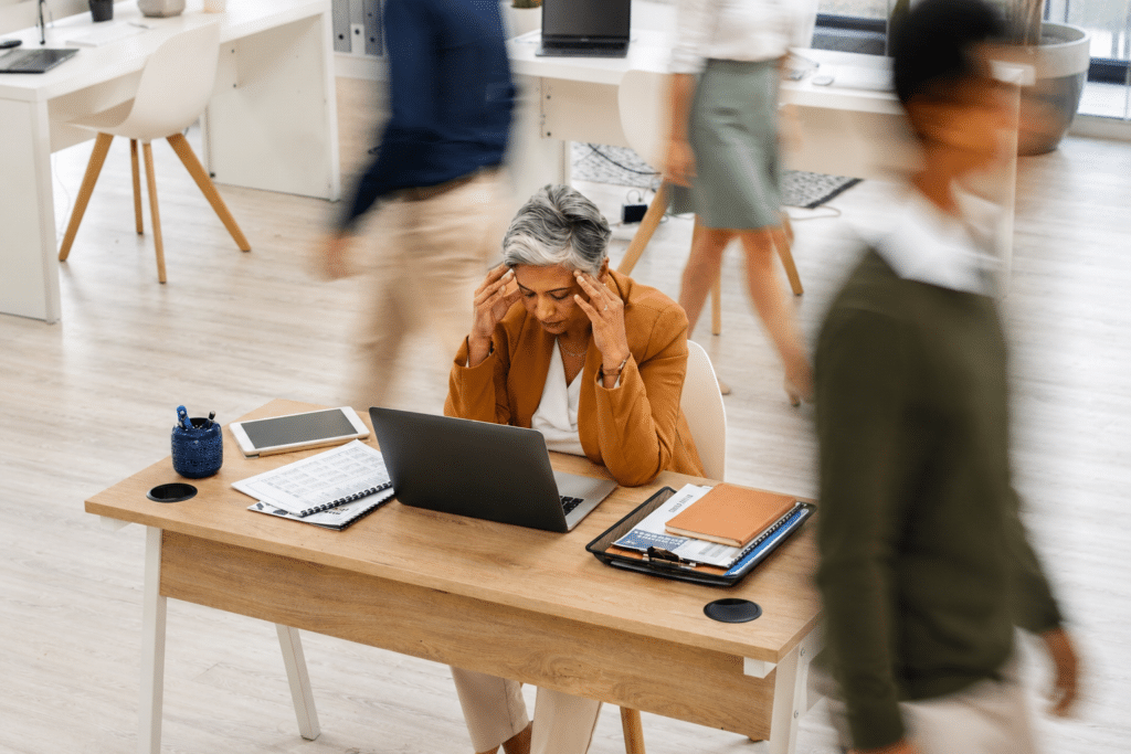 A professional woman sits at her light wood desk in a busy office environment