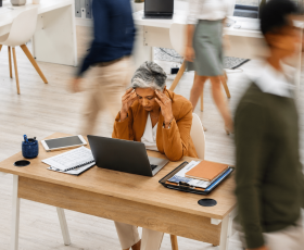 A professional woman sits at her light wood desk in a busy office environment