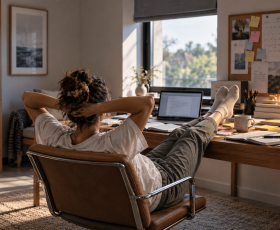 A woman reclines in her home office chair with her feet on a cluttered desk, surrounded by unfinished paperwork, notebooks, and a laptop, illustrating ADHD procrastination and difficulty starting tasks.